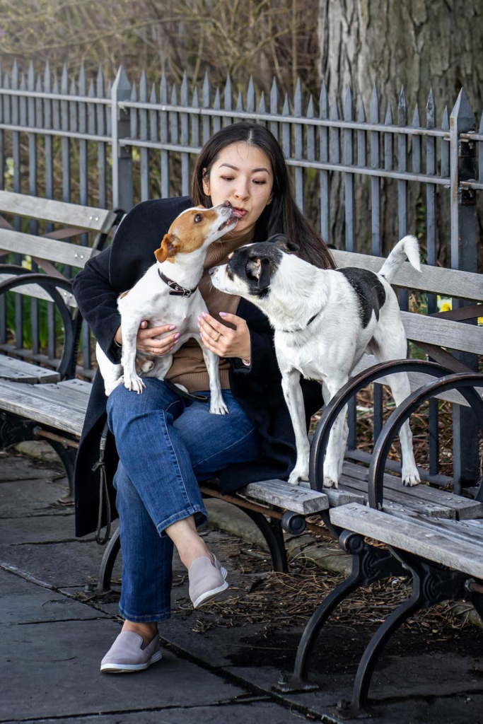 Woman sits on a park bench with two dogs; one playfully licks her face while the other looks on.
