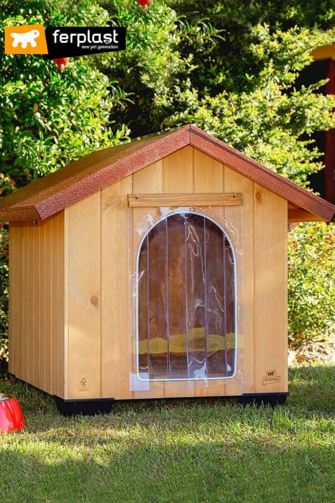 A wooden dog house with a slanted asphalt roof and a clear plastic flap door.