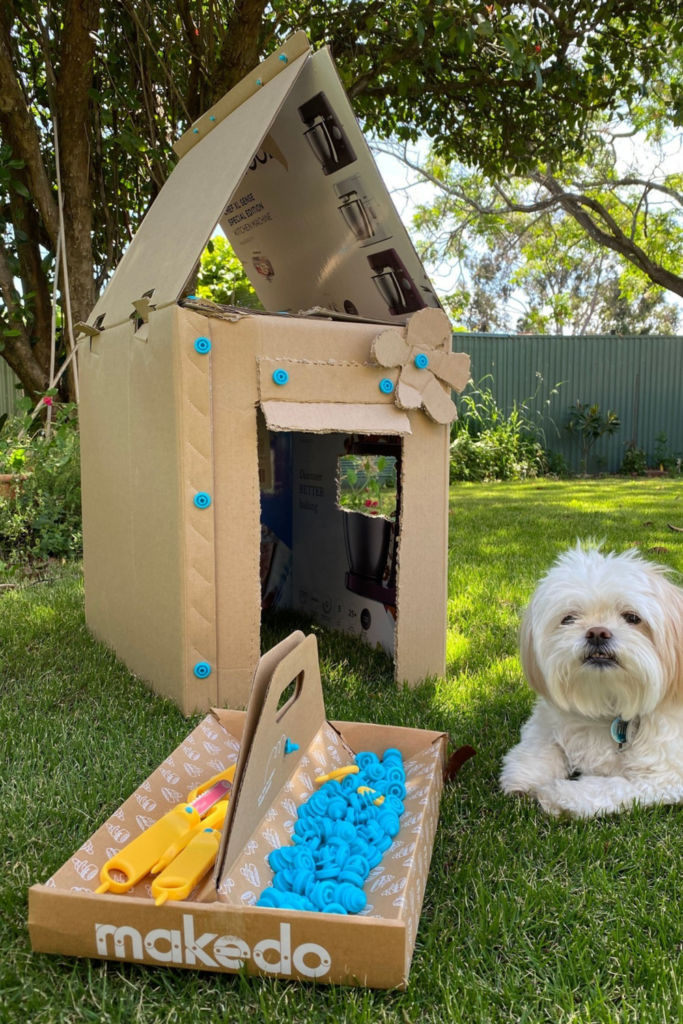 A creative dog kennel made of cardboard decorated to look like a castle. A white dog sitting beside the kennel.