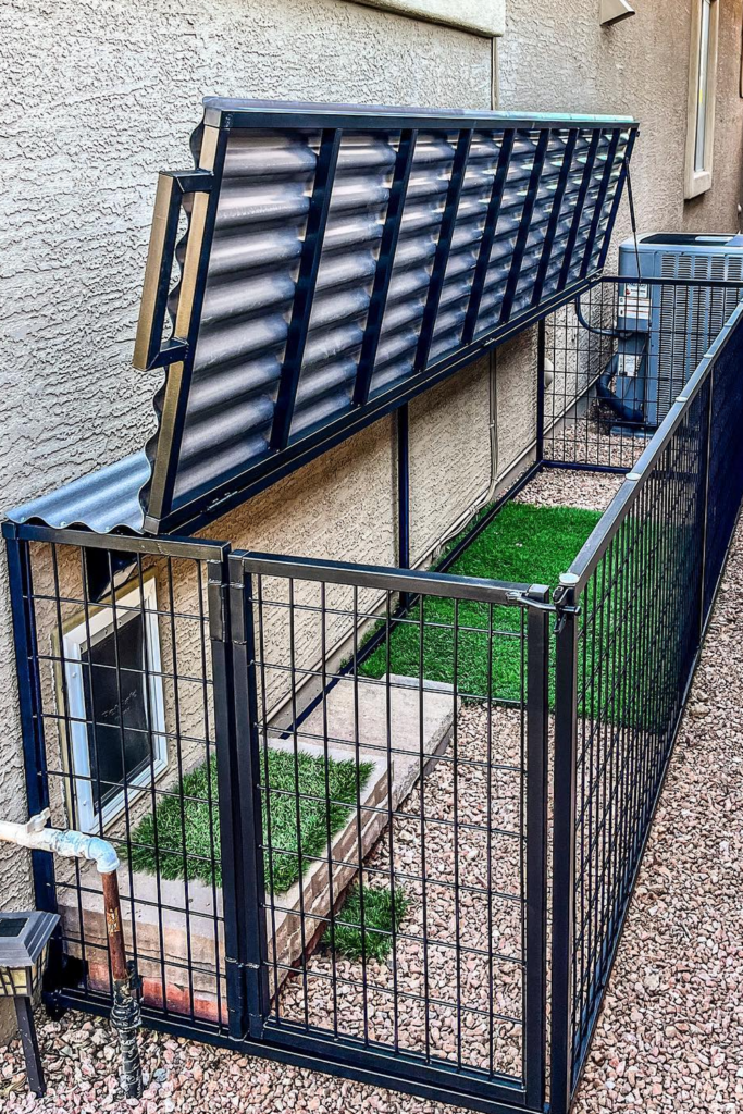A large wire dog kennel with a metal roof. The kennel has a secure gate and a green grass carpet visible inside.
