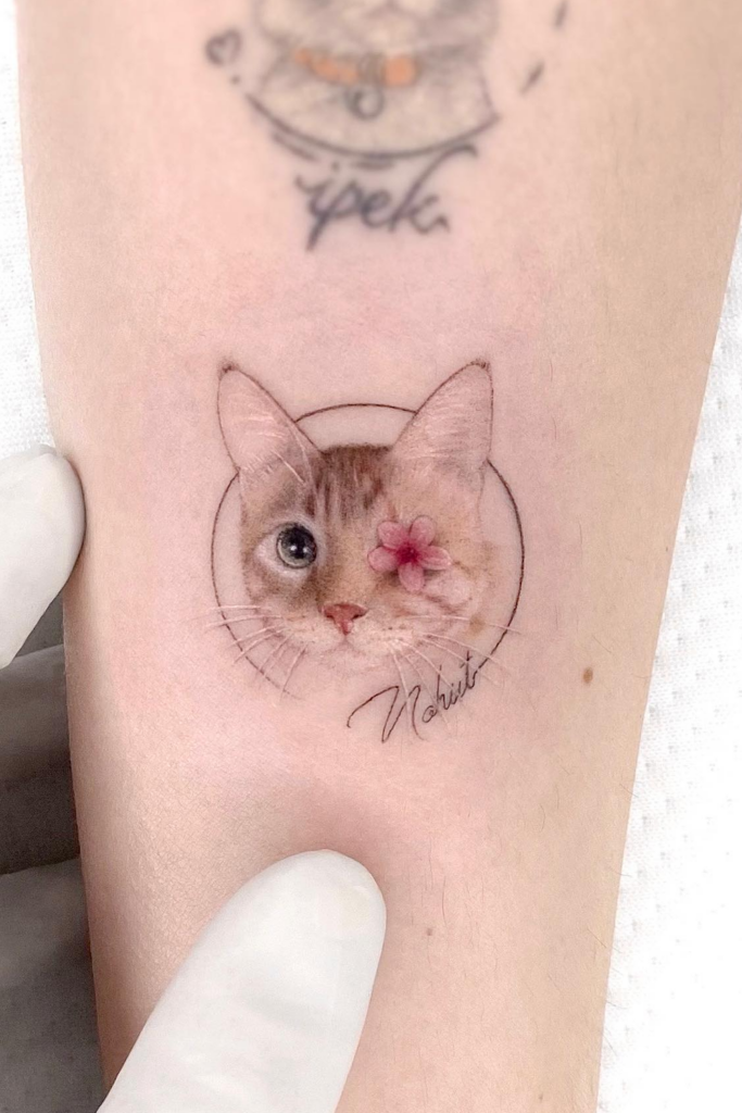 Tattoo of a one-eyed cat, the missing eye delicately covered by pink flower petals.