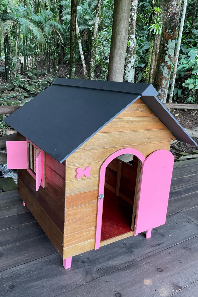 A small wooden dog house painted brown and pink with a black roof sits on a wooden deck.