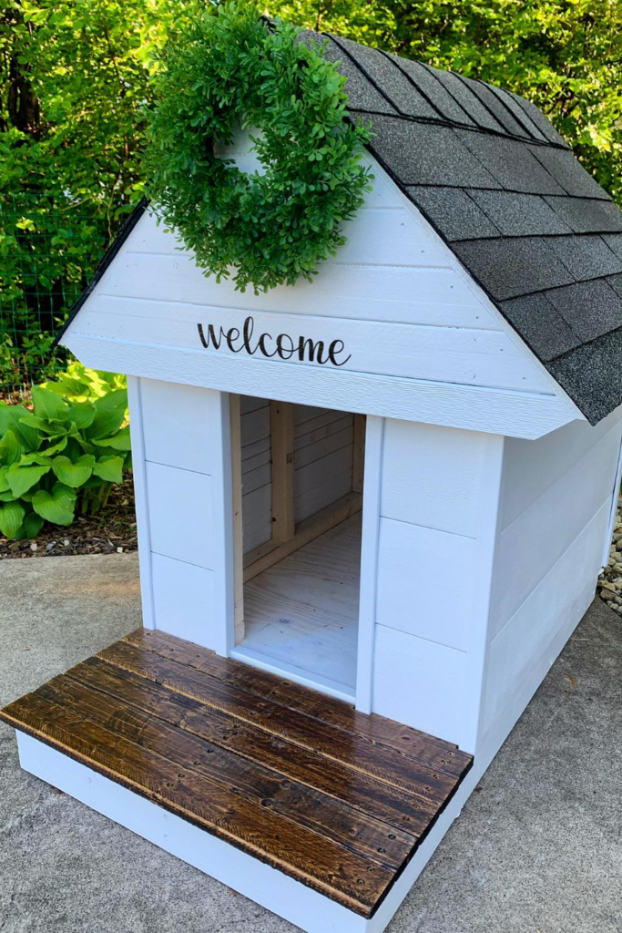A white dog house with a dark grey roof and a wreath on the roof. The door of the dog house is decorated with a welcome sign.