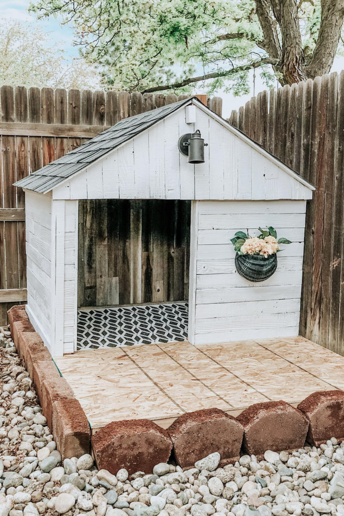 A small wooden dog house painted white with a black roof sits on a gravel backyard. There is a potted flower next to the door.