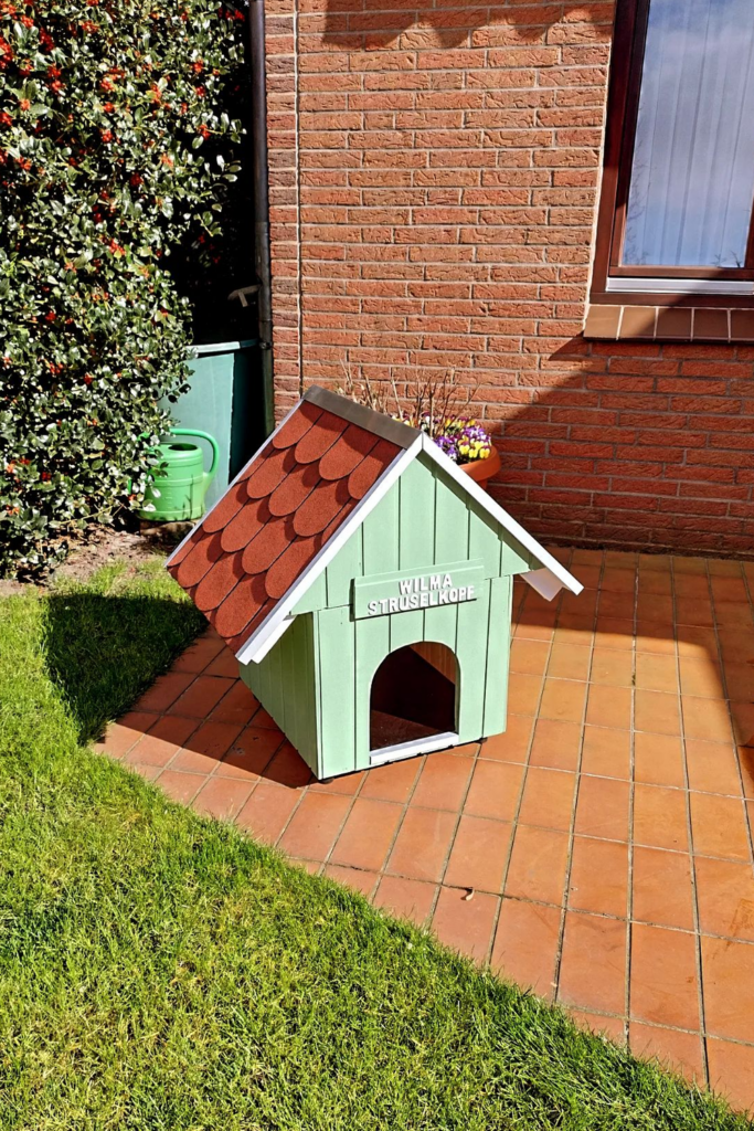 A green dog house with a red roof rests on a beautiful backyard.