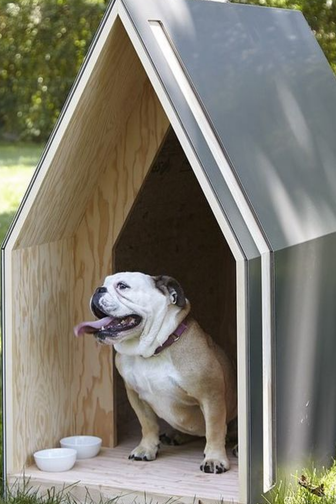 A small, modern dog house with an inverted 'V' shaped roof. A bulldog sits along the doorway
